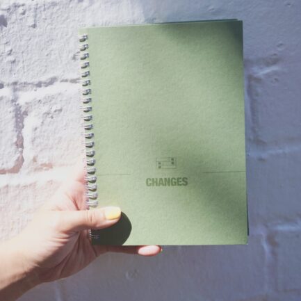 Green manuscript notebook being held in someone's hand, with the Changes logo on the front of it, glistening in the sun.