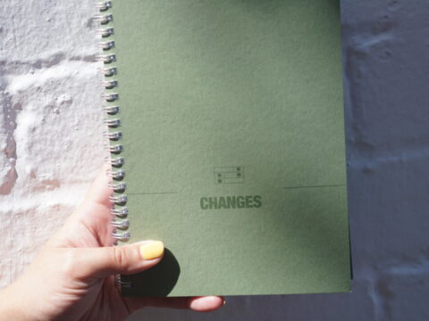 Green manuscript notebook being held in someone's hand, with the Changes logo on the front of it, glistening in the sun.