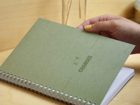 A green manuscript notebook being opened by someone's hand, with the Changes logo on the front of it.