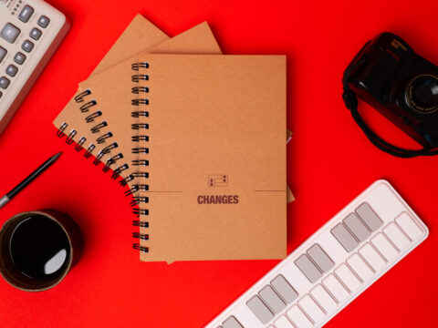 A brown music manuscript notebook with wirobinding and the Changes logo on the front, surrounded by a ceramic cup of coffee, a pencil, a midi keyboard, a synth and a camera.